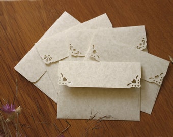 May include: A stack of six beige envelopes with delicate lace-like cutouts around the edges. The envelopes are arranged in a staggered pattern on a wooden surface.