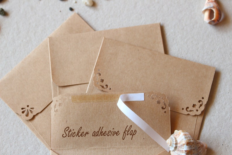May include: Kraft paper envelopes with decorative corner cutouts and a sticker adhesive flap. The text "Sticker adhesive flap" is printed on the front of one envelope. A white ribbon is attached to the flap. Seashells and small stones are in the background.