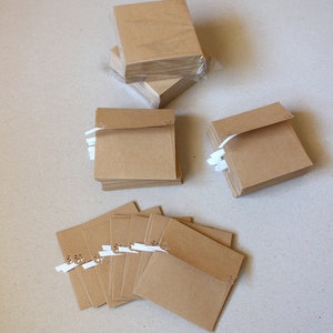 May include: Multiple stacks of small, square kraft paper cards. Some are wrapped in clear plastic, others are fanned out. Each card features a decorative cut-out and a white ribbon. The cards are arranged on a neutral surface.