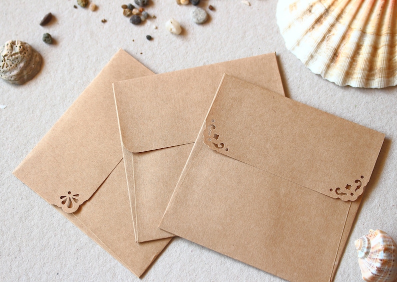 May include: Three brown paper envelopes with decorative corner cutouts, arranged on a light surface. Seashells and small pebbles are scattered around the envelopes. The envelopes are a natural, neutral colour.