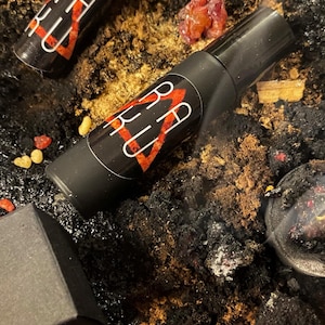 May include: Two black roller bottles with red and white labels that say "BELLU" on a dark background with orange and red pieces of resin.