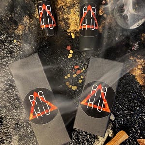 May include: Two black boxes with a red triangle logo that says "RAKU" and two black roller bottles with the same logo. The bottles and boxes are on a black surface with gold and red accents.