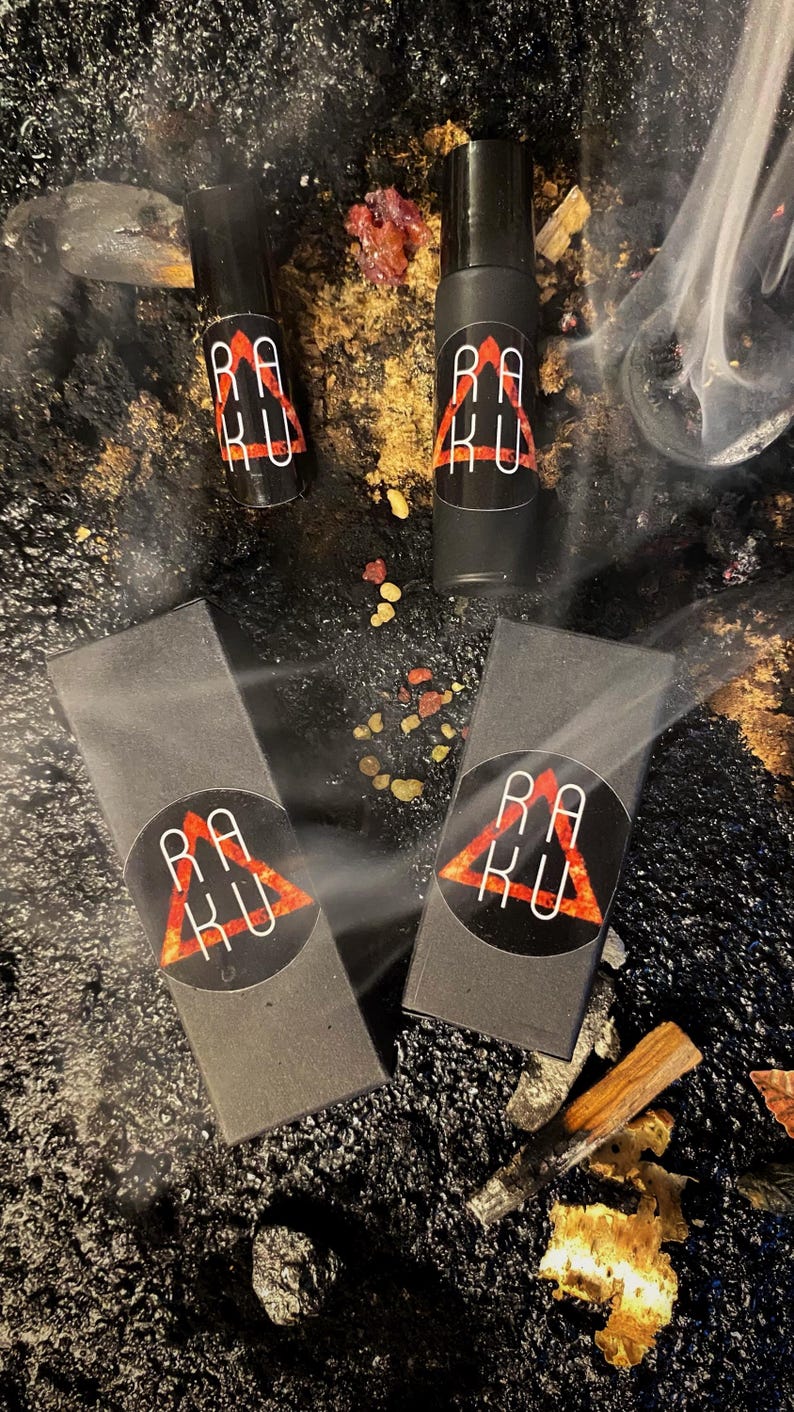 May include: Two black boxes with a red triangle logo that says "RAKU" and two black roller bottles with the same logo. The bottles and boxes are on a black surface with gold and red accents.
