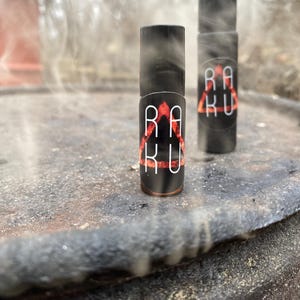 May include: Two small black bottles with red and white labels that say "RAKU" in a triangular design. The bottles are sitting on a rusty surface.