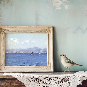 A framed seascape painting with a sailboat sits on a rustic shelf, alongside a small bird. The scene is calm, with a lace cloth adding a vintage touch.