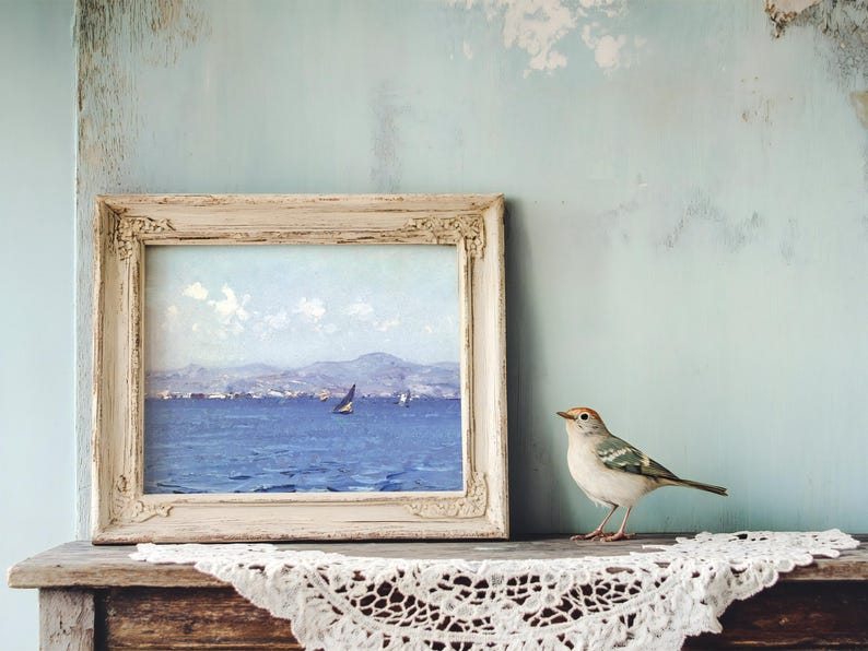 A framed seascape painting with a sailboat sits on a rustic shelf, alongside a small bird. The scene is calm, with a lace cloth adding a vintage touch.