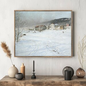 A framed landscape painting hangs on a white wall above a wooden console with vases and a candle. The painting depicts a snowy village scene with houses and trees, conveying a serene winter atmosphere.