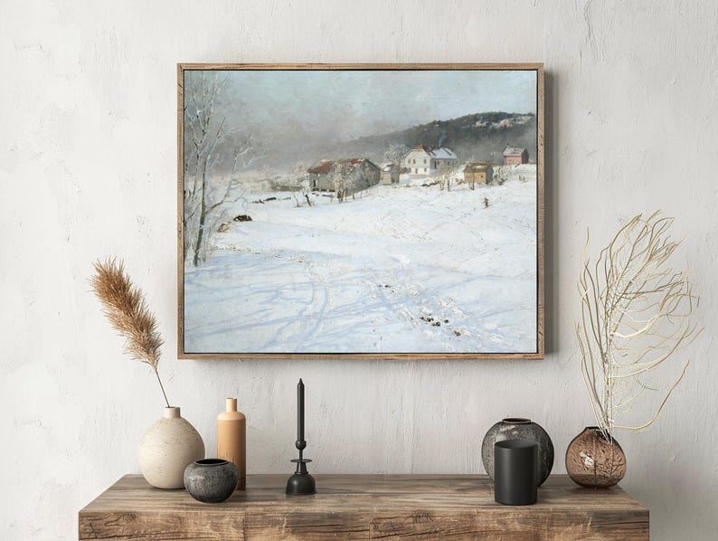 A framed landscape painting hangs on a white wall above a wooden console with vases and a candle. The painting depicts a snowy village scene with houses and trees, conveying a serene winter atmosphere.