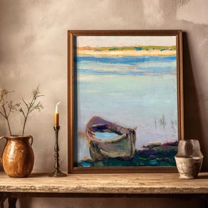 A framed painting of a boat on calm water sits on a wooden table. A ceramic jug and candlestick with dried plants are beside it, creating a rustic, serene atmosphere.