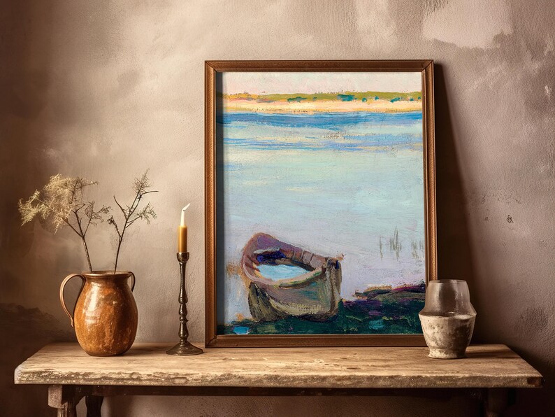 A framed painting of a boat on calm water sits on a wooden table. A ceramic jug and candlestick with dried plants are beside it, creating a rustic, serene atmosphere.