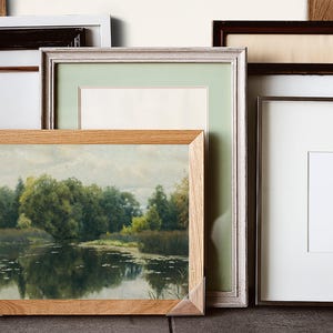 A collection of framed art and photographs. The frames vary in style, including wood, white, and dark brown. One frame contains a landscape painting with trees and water. The other frames are empty with white or light green matting.