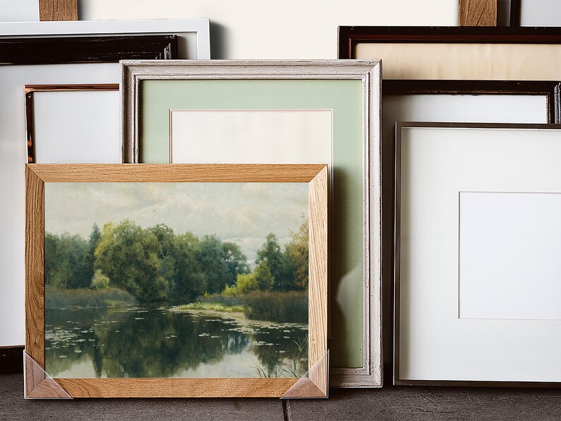 A collection of framed art and photographs. The frames vary in style, including wood, white, and dark brown. One frame contains a landscape painting with trees and water. The other frames are empty with white or light green matting.