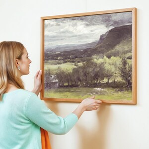 A woman with blonde hair and a turquoise sweater adjusts a framed landscape painting on a white wall, depicting a serene, cloudy countryside.