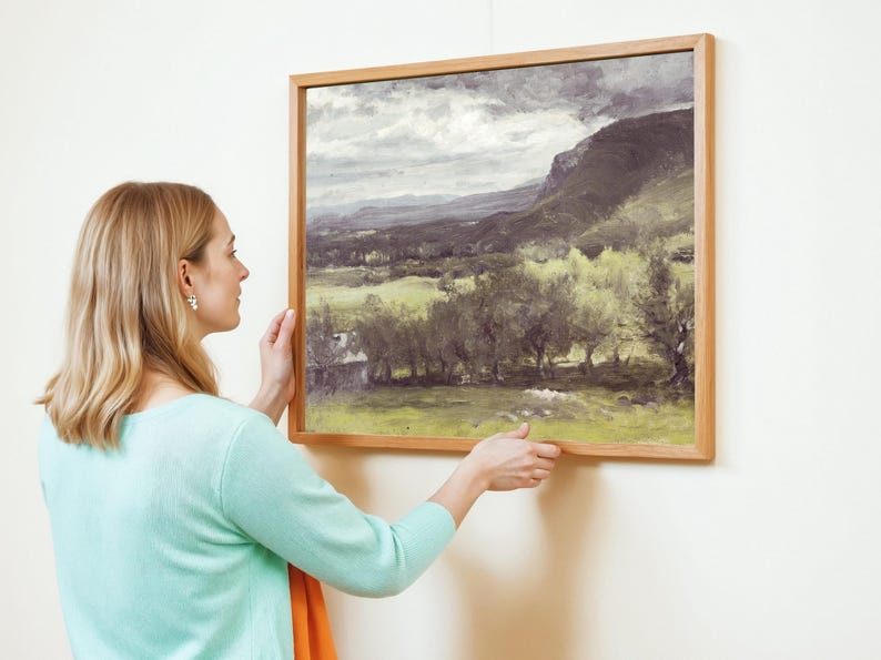 A woman with blonde hair and a turquoise sweater adjusts a framed landscape painting on a white wall, depicting a serene, cloudy countryside.
