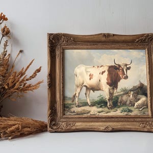 A framed painting of a cow and sheep in a pastoral setting with a cloudy sky. Beside it, dried flowers and grasses in a rustic vase on a wooden surface.