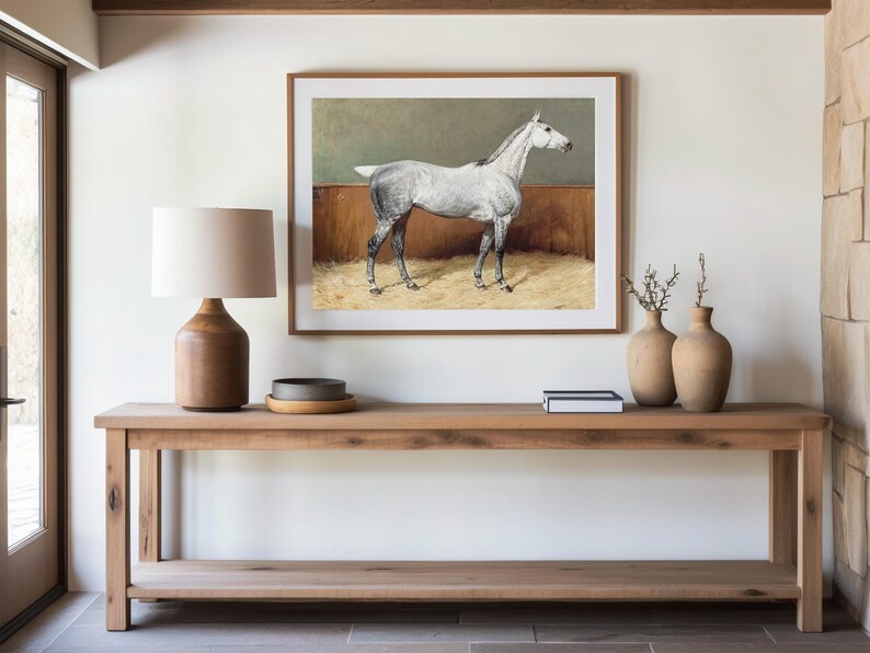 A minimalist entryway features a wooden console table with a lamp, small tray, books, and vases. A framed horse painting hangs above, creating a serene atmosphere.