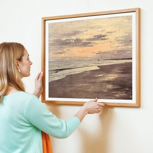 A woman in a turquoise sweater adjusts a framed painting of a serene beach sunset. The painting features soft, muted tones, evoking calm and tranquility.