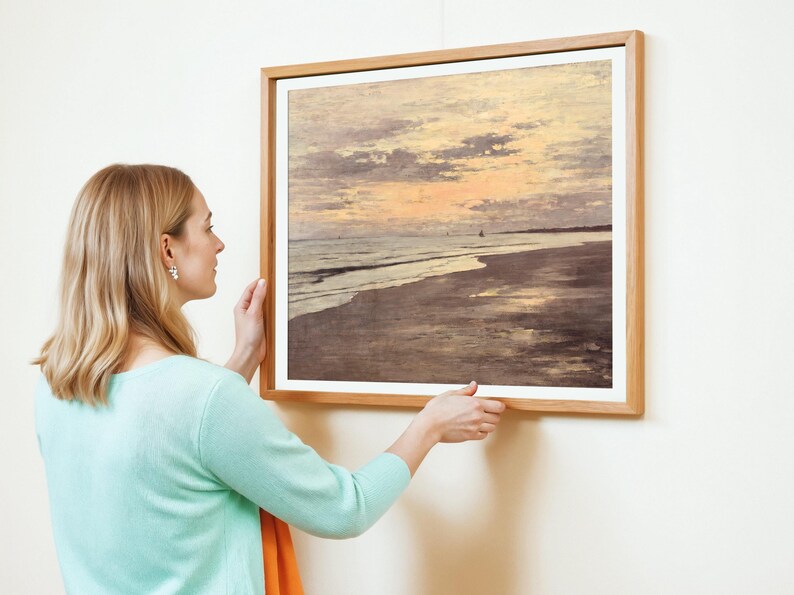 A woman in a turquoise sweater adjusts a framed painting of a serene beach sunset. The painting features soft, muted tones, evoking calm and tranquility.