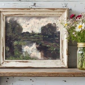 A rustic framed painting of a serene river scene sits on a wooden shelf. Next to it, a glass jar holds vibrant daisies and wildflowers, creating a cozy, peaceful ambiance.