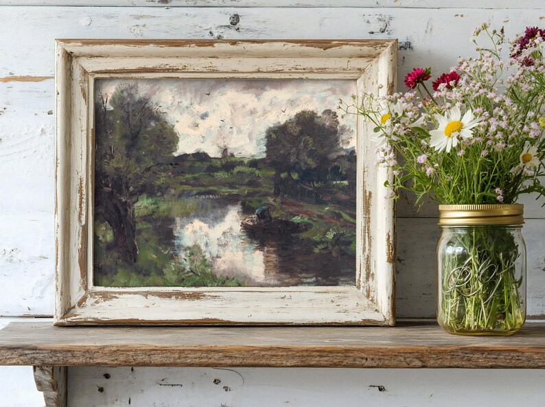 A rustic framed painting of a serene river scene sits on a wooden shelf. Next to it, a glass jar holds vibrant daisies and wildflowers, creating a cozy, peaceful ambiance.