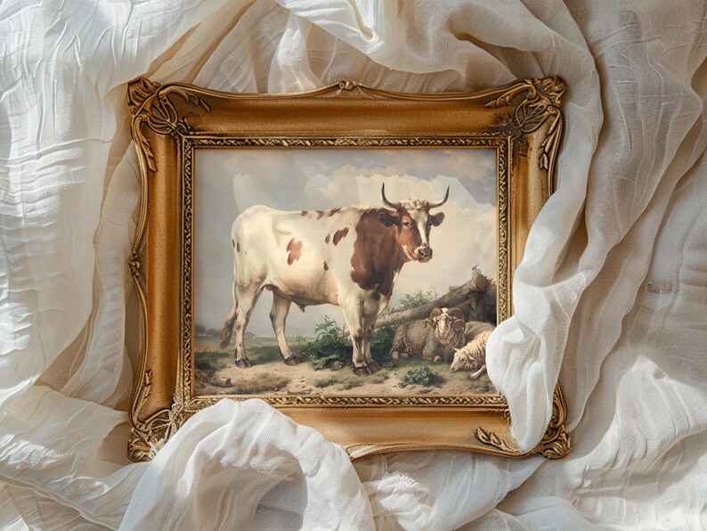 A painting of a cow and sheep in a pastoral scene is framed elaborately in gold. The frame rests on soft, crumpled white fabric, creating a serene and classic atmosphere.