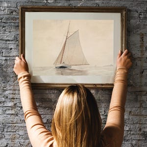 A woman hangs a framed watercolor painting of a sailboat on a brick wall. The sailboat is white with a brown mast and sails. The painting is in a simple wooden frame.