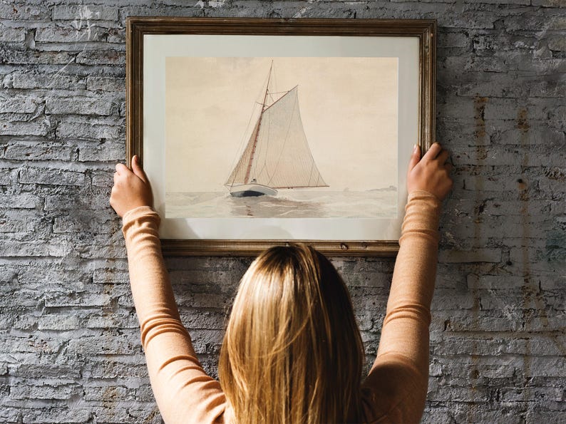 A woman hangs a framed watercolor painting of a sailboat on a brick wall. The sailboat is white with a brown mast and sails. The painting is in a simple wooden frame.