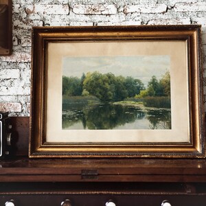 A framed landscape painting with a gold ornate frame. The artwork depicts a serene lake scene with trees and reflections in the water. The painting is displayed on a dark wooden surface.