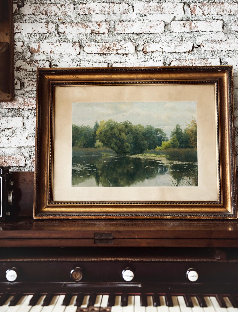 A framed landscape painting with a gold ornate frame. The artwork depicts a serene lake scene with trees and reflections in the water. The painting is displayed on a dark wooden surface.