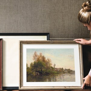 A framed landscape painting with a gold-colored frame. The artwork depicts a river scene with trees and a small village in the background. The painting is being held by a person.