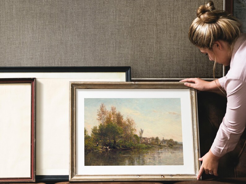 A framed landscape painting with a gold-colored frame. The artwork depicts a river scene with trees and a small village in the background. The painting is being held by a person.