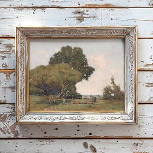 A framed landscape painting leaning against a weathered white wooden wall, depicting a scene with large trees, a grassy field, and a person sitting on the ground under the trees on a partly cloudy day.
