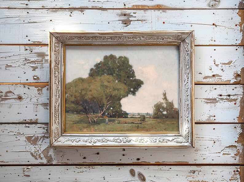 A framed landscape painting leaning against a weathered white wooden wall, depicting a scene with large trees, a grassy field, and a person sitting on the ground under the trees on a partly cloudy day.