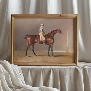 Framed painting of a jockey on a brown horse, set against a muted background. The frame rests on a draped fabric, conveying elegance and calm.