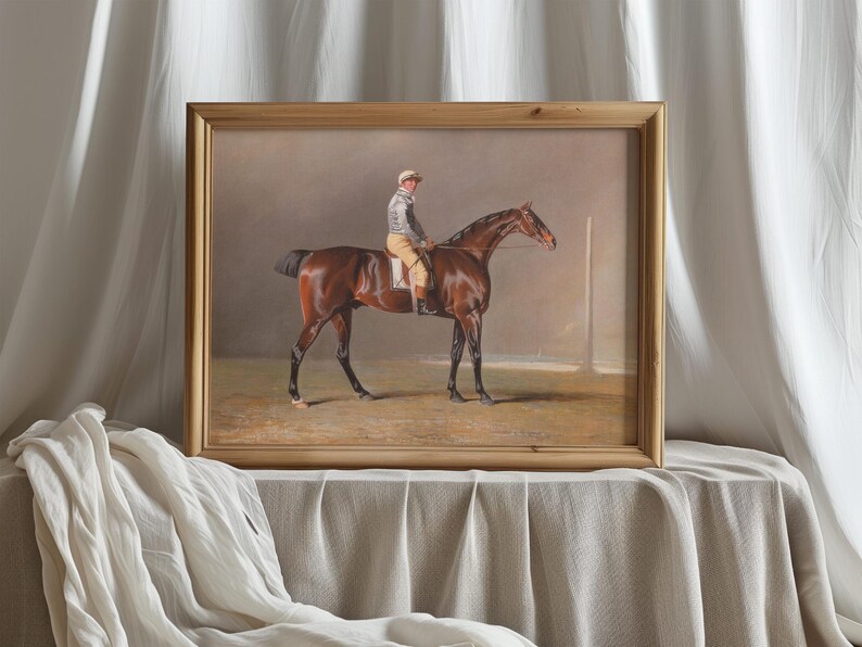 Framed painting of a jockey on a brown horse, set against a muted background. The frame rests on a draped fabric, conveying elegance and calm.