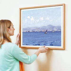 A woman in a mint green sweater hangs a framed seascape painting on a white wall. The artwork depicts a sailboat on a calm blue sea under a clear sky.