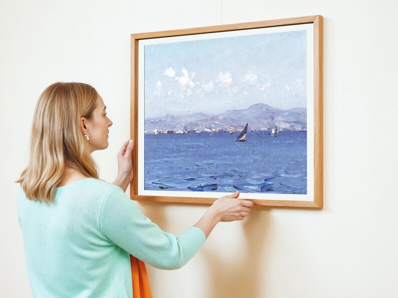A woman in a mint green sweater hangs a framed seascape painting on a white wall. The artwork depicts a sailboat on a calm blue sea under a clear sky.