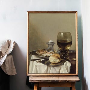A framed still life painting on an easel shows wine, bread, and delicacies on a white tablecloth. The setting is calm and elegant, with muted tones.