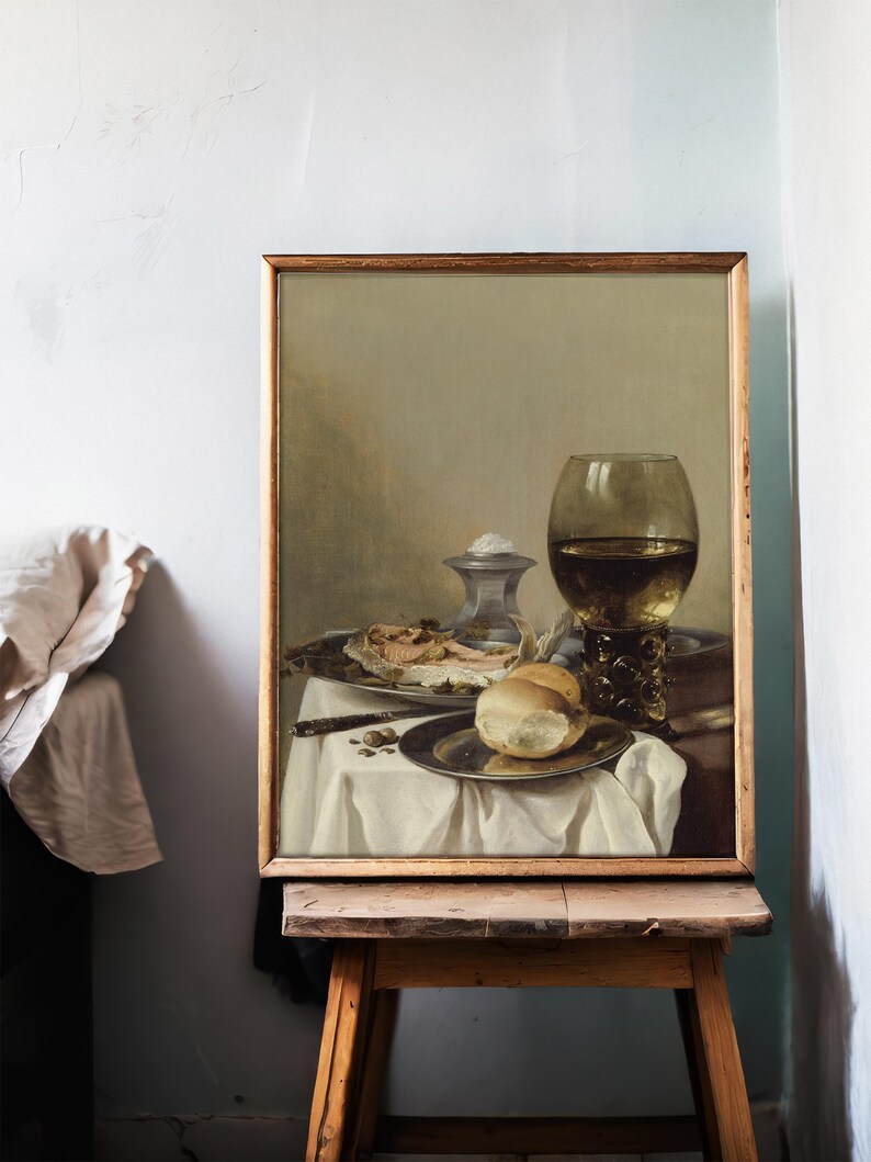 A framed still life painting on an easel shows wine, bread, and delicacies on a white tablecloth. The setting is calm and elegant, with muted tones.