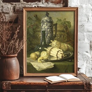 Framed still-life painting on a rustic table against a textured brick wall. Includes asparagus, lemons, and a jug, with an open book nearby. Cozy, vintage feel.