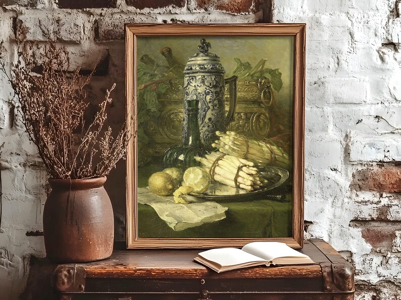Framed still-life painting on a rustic table against a textured brick wall. Includes asparagus, lemons, and a jug, with an open book nearby. Cozy, vintage feel.