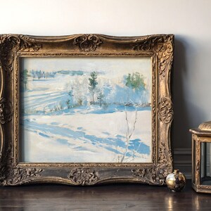 An ornate framed painting of a snowy landscape rests against a wall. A vintage lantern and small metallic sphere sit beside it on the wooden floor.