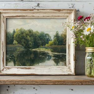 A rustic framed painting of a serene river scene with lush greenery sits on a wooden shelf. Beside it, a mason jar holds wildflowers, adding a touch of nature.