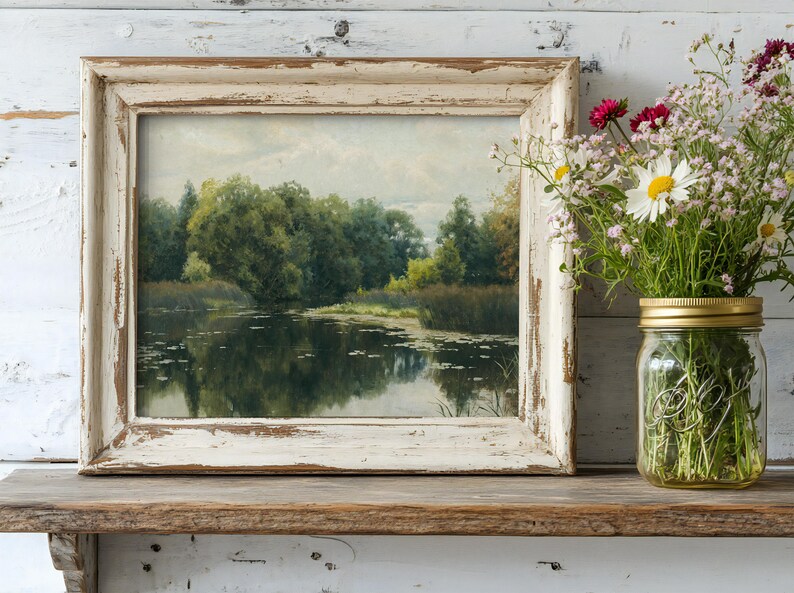 A rustic framed painting of a serene river scene with lush greenery sits on a wooden shelf. Beside it, a mason jar holds wildflowers, adding a touch of nature.