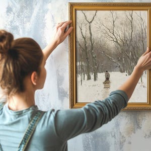 A woman in a blue shirt is hanging a framed winter landscape painting on a textured wall. The painting depicts a snowy path lined with bare trees, conveying a tranquil mood.