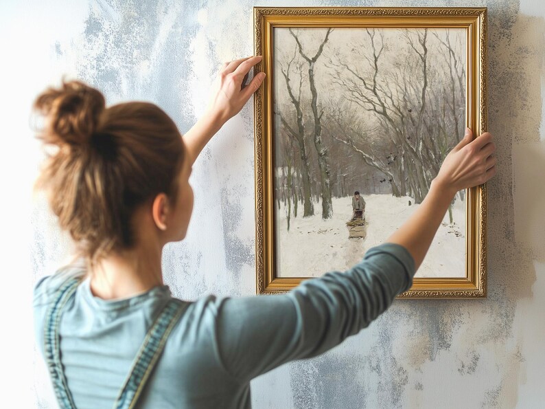 A woman in a blue shirt is hanging a framed winter landscape painting on a textured wall. The painting depicts a snowy path lined with bare trees, conveying a tranquil mood.