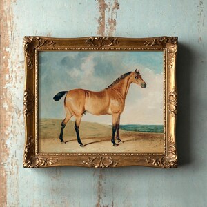 A framed painting of a horse standing in a field is mounted on a textured, weathered blue wall. The ornate golden frame adds classic elegance.
