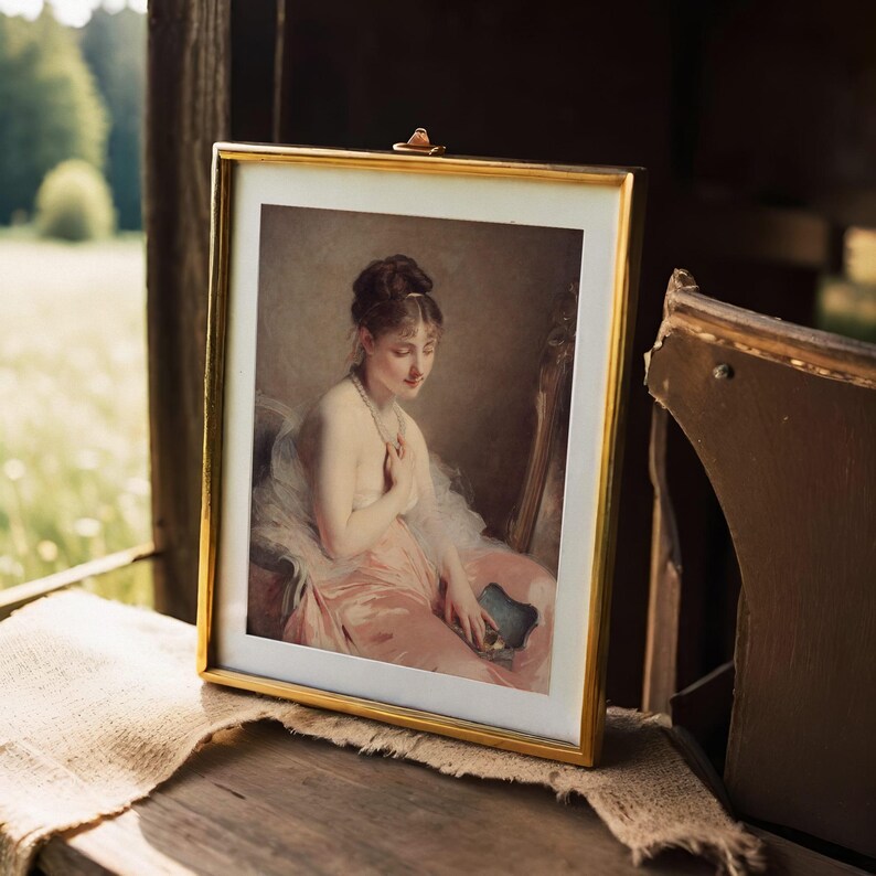 Framed painting of a seated woman in a pink dress, holding a fan, set on a wooden table with a cloth cover, against a soft-focus outdoor background.