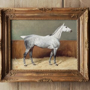 Framed painting of a white horse standing on hay, with a wooden wall background. Ornate gold frame contrasts with the rustic setting.