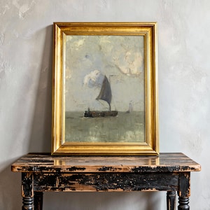 A framed painting of a sailing boat on a calm sea with cloudy skies is placed on a rustic wooden table against a textured gray wall, evoking serenity.